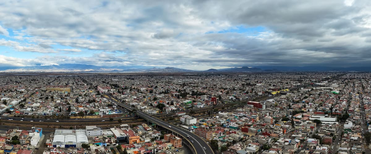A sprawling urban landscape with a dense grid of buildings, a prominent highway, and distant mountains under a cloudy sky.