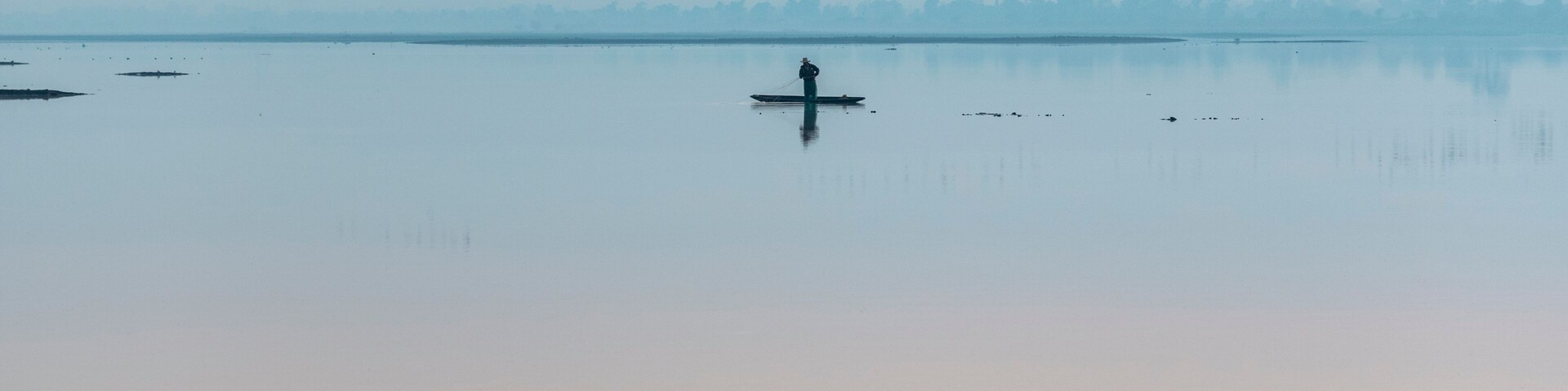 Un lago tranquilo con una persona remando en kayak, rodeado por un bosque brumoso y una bandada de pájaros a lo lejos.