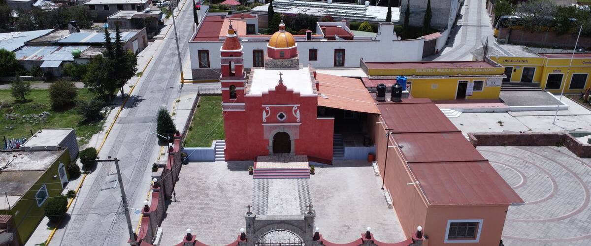 A church with a red facade and a yellow dome, surrounded by a paved square and a curved road.