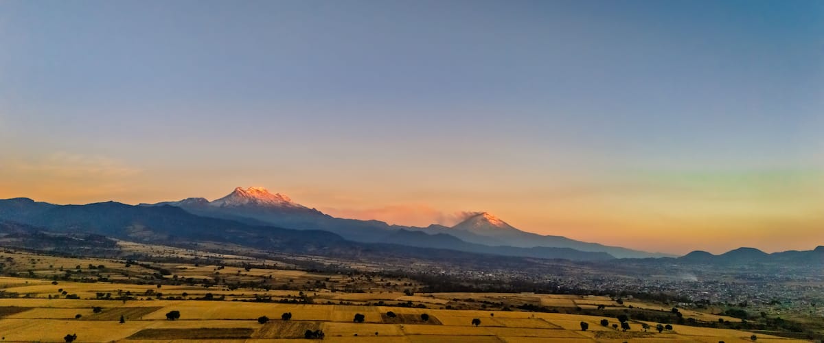A mountain range with a prominent peak, a valley with fields, and a distant cityscape.
