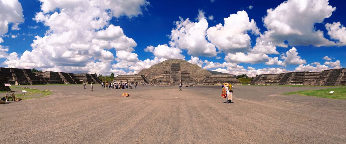 A large ancient pyramid with a flat top, surrounded by smaller structures and a clear blue sky with clouds.