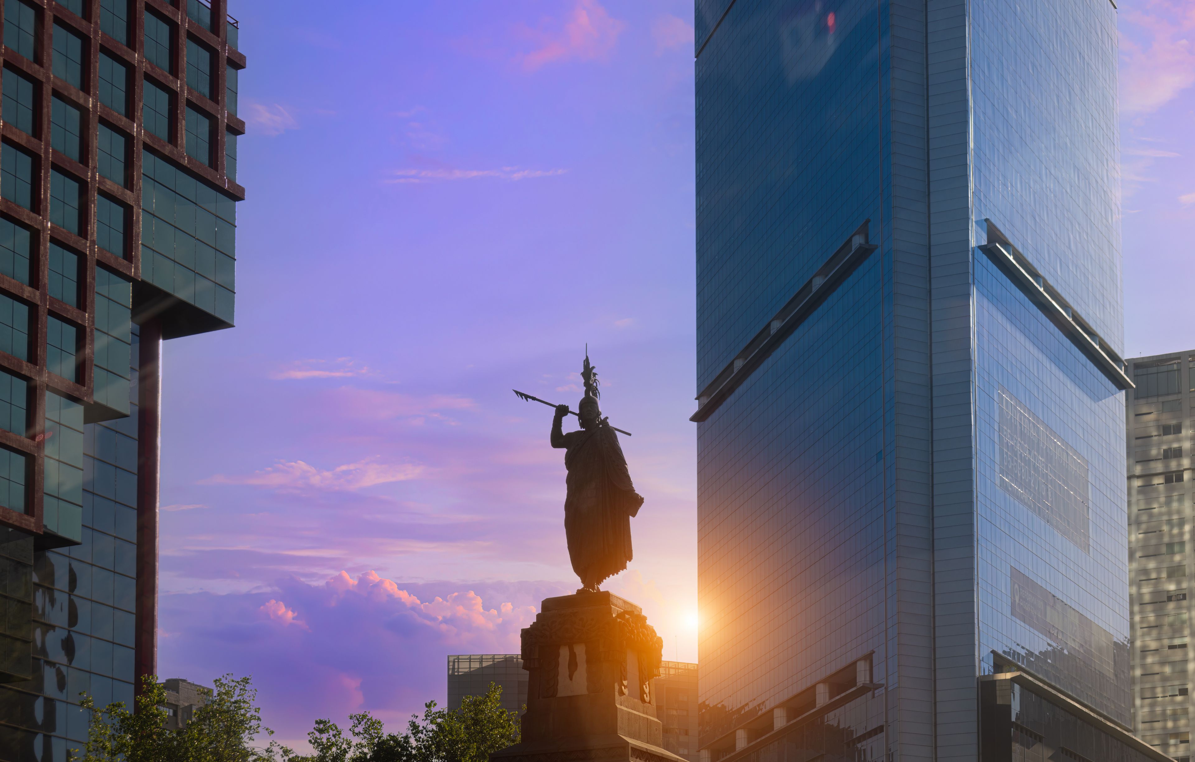 Uma estátua em ambiente urbano, com edifícios modernos e um pôr do sol ao fundo.