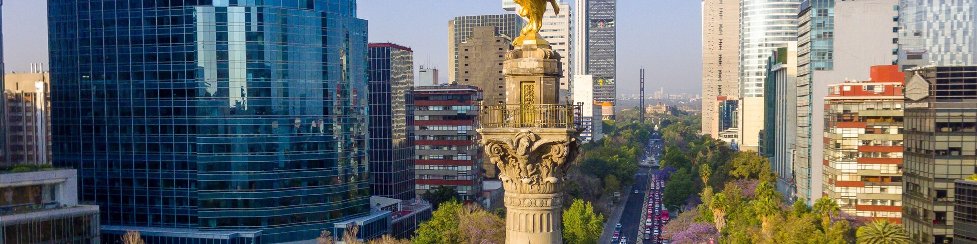 Un paisaje urbano con una prominente estatua dorada sobre una columna, modernos edificios altos y una calle concurrida llena de coches.