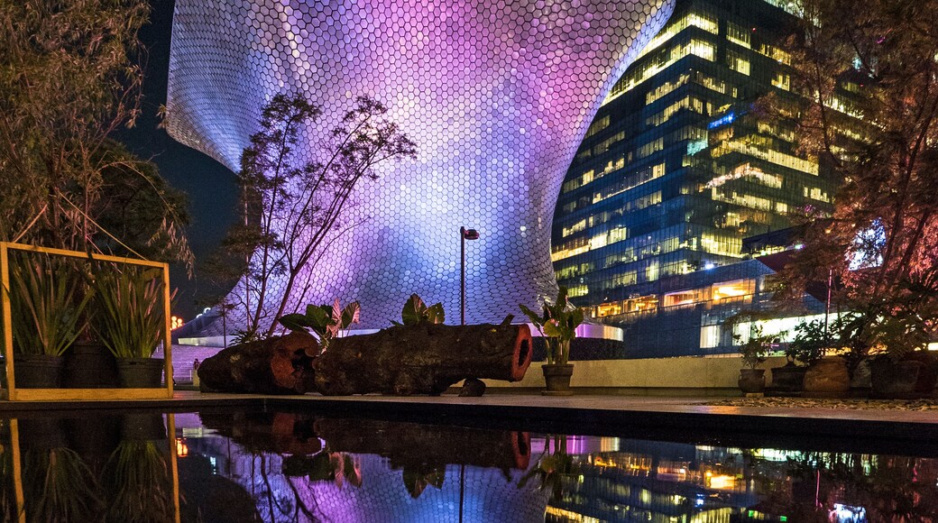 Un edificio moderno con fachada hexagonal iluminada durante la noche, reflejándose en una superficie acuática tranquila.