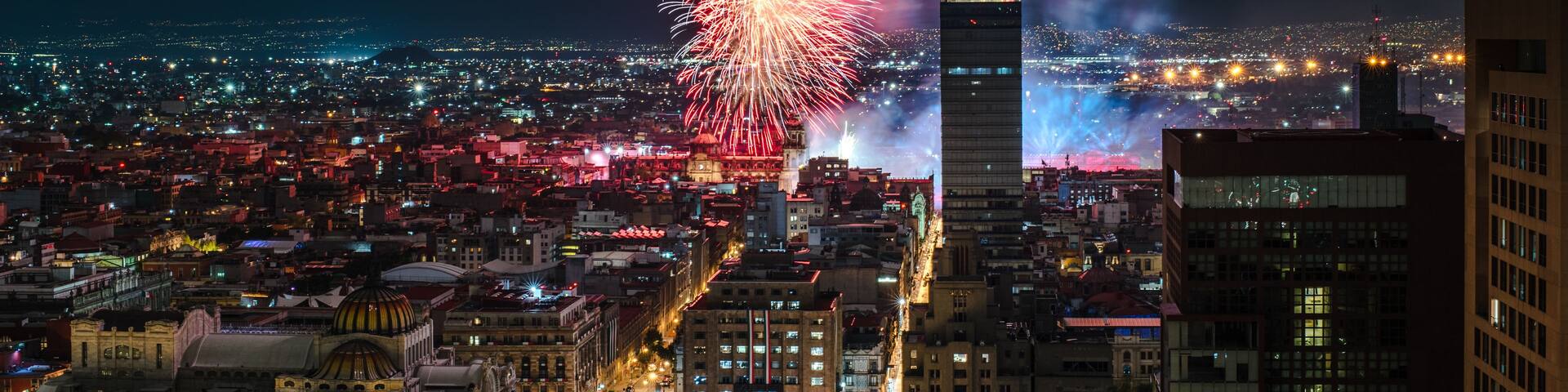 A cityscape at night with fireworks exploding in the sky above a prominent skyscraper.