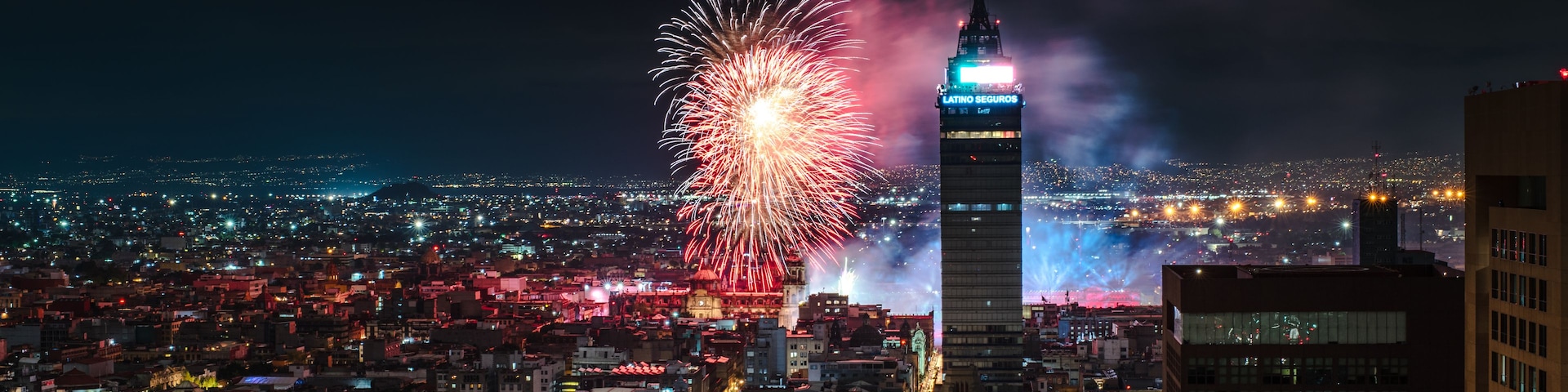 A cityscape at night with fireworks exploding in the sky above a prominent skyscraper.