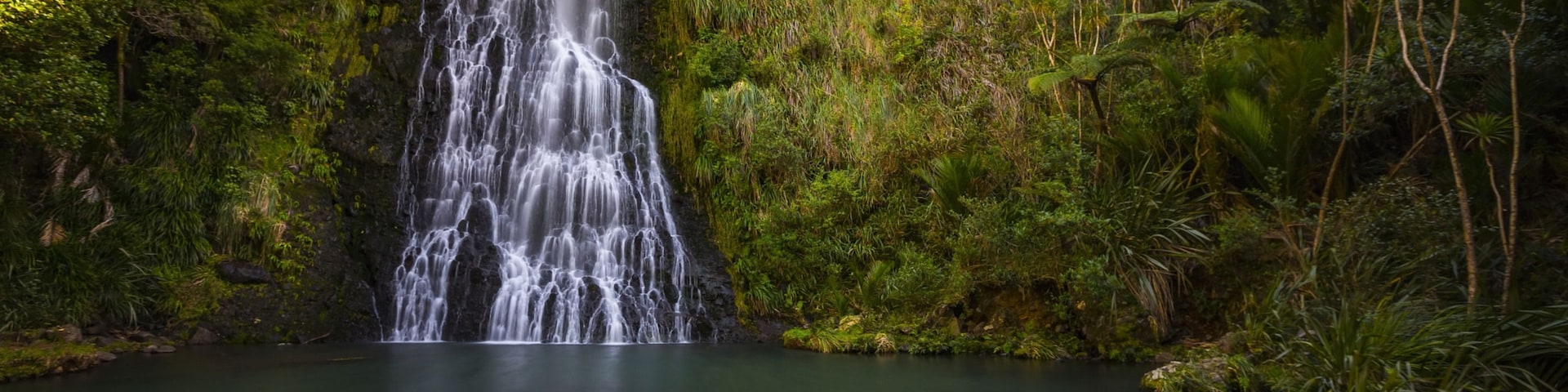 Uma cascata despencando em uma piscina tranquila, cercada por vegetação exuberante.