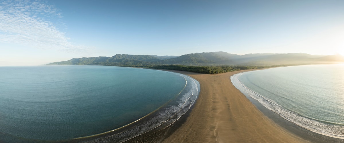 A coastal landscape with a curved beach, calm sea, and distant mountains.
