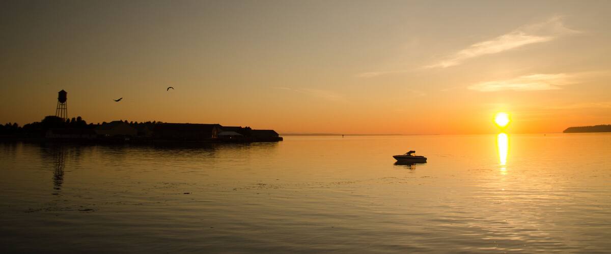 A sunset over a calm body of water with a small boat and distant buildings silhouetted against the sky.
