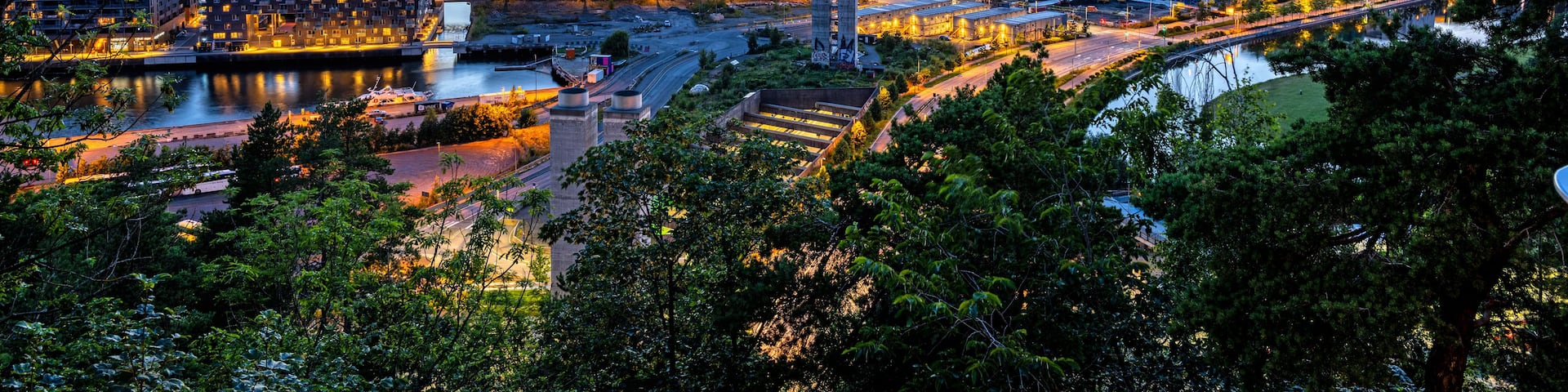 A cityscape at dusk with a river, buildings, and mountains in the background.