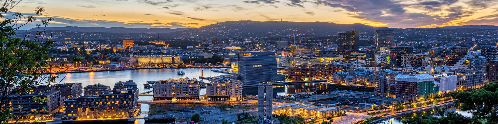A cityscape at dusk with a river, buildings, and mountains in the background.