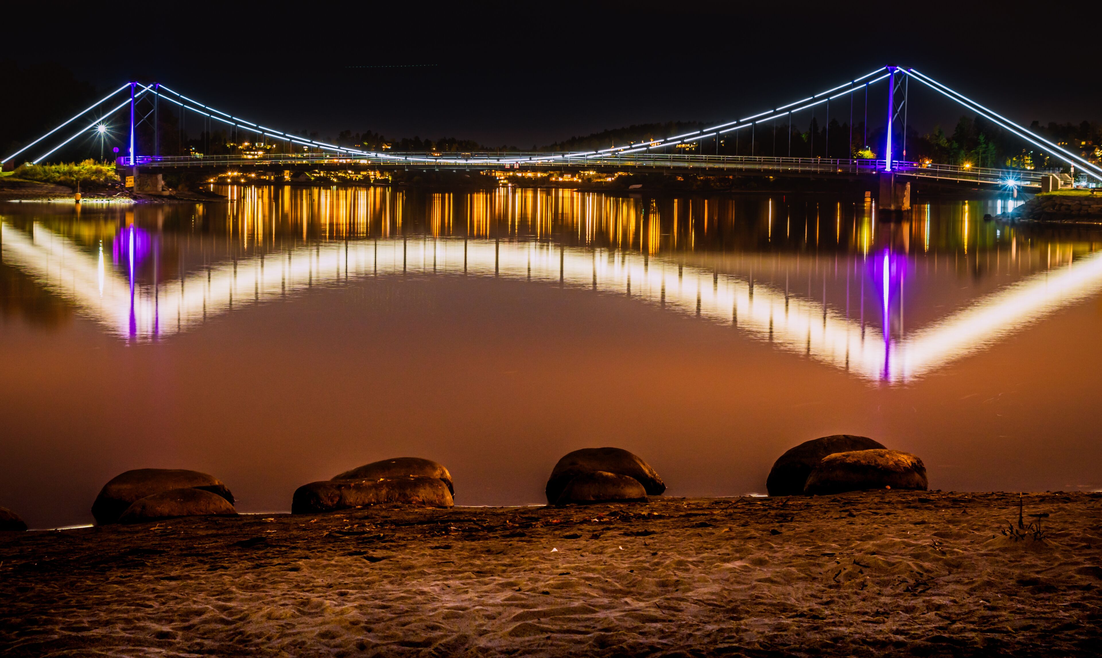 A lit bridge at night reflecting on calm water with rocks in the foreground.