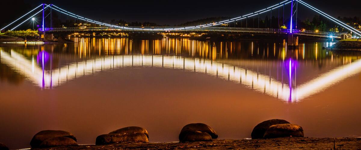 A lit bridge at night reflecting on calm water with rocks in the foreground.