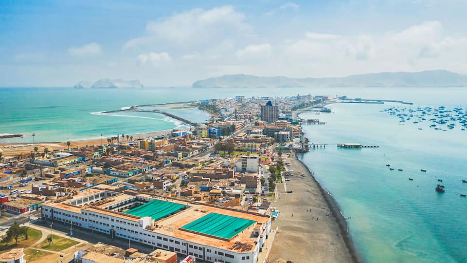 A coastal cityscape with a prominent white building with green roofs, a beach, and a marina with numerous boats.