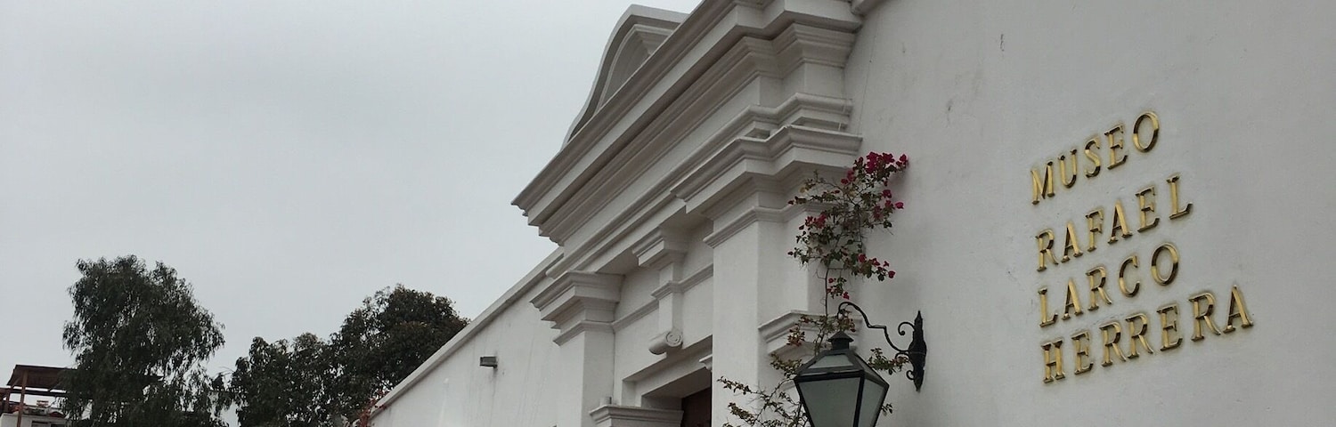 A building with a white facade and a red flag. The building has a sign that reads "Museo Rafael Larc Herrera". There are potted plants and flowers around the building.