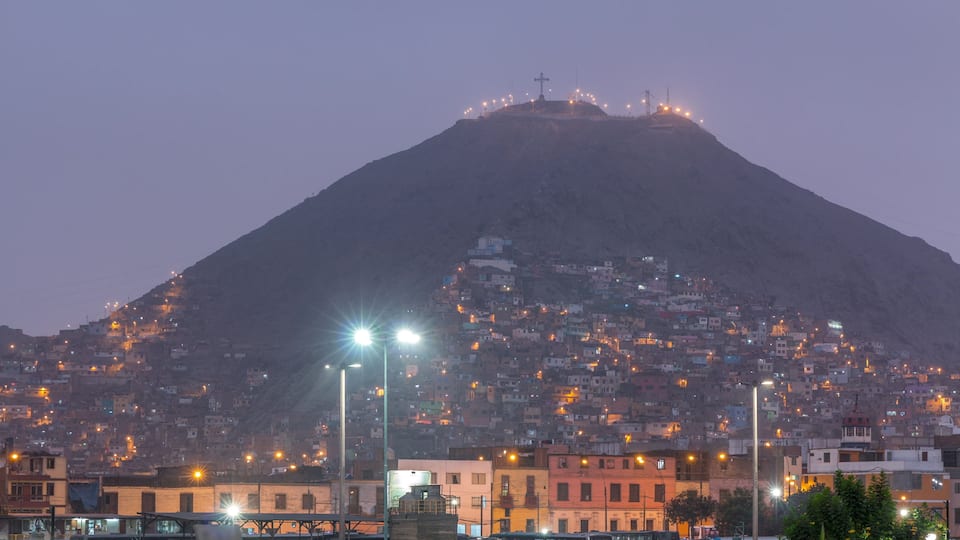 Una montaña con una cruz en la cima, una ladera densamente poblada debajo y farolas iluminando la zona.