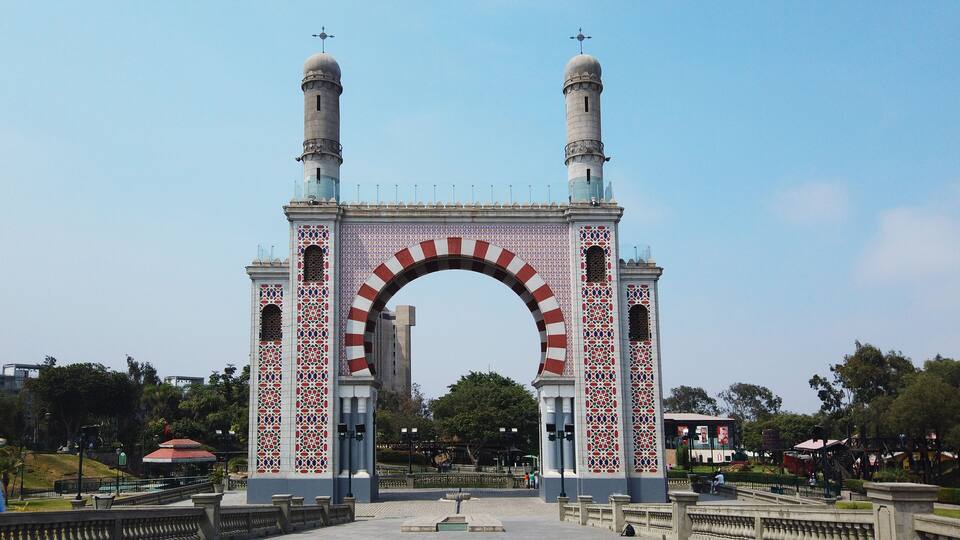 A large, ornate archway with intricate tile work and two towers, set in a park with trees and a fountain.
