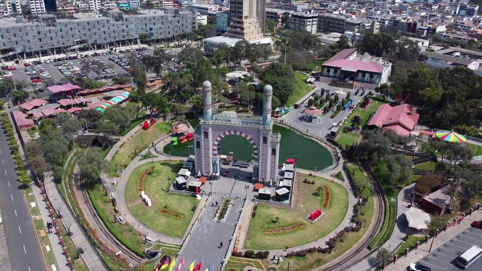 An aerial view of a park with a central fountain, surrounding pathways, and a large building with a red roof.