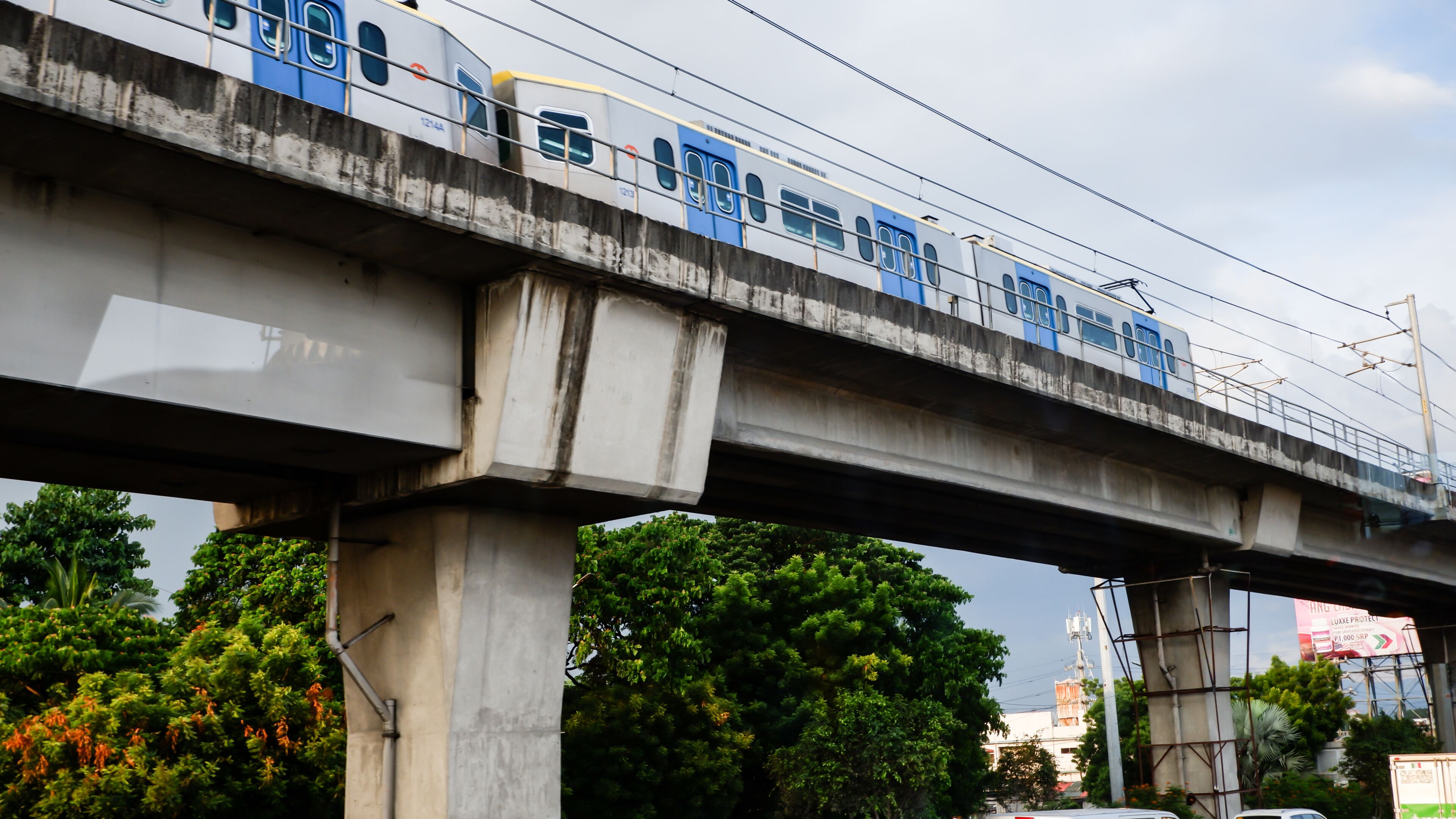 A train on an elevated track with concrete pillars, greenery below, and a billboard in the background.