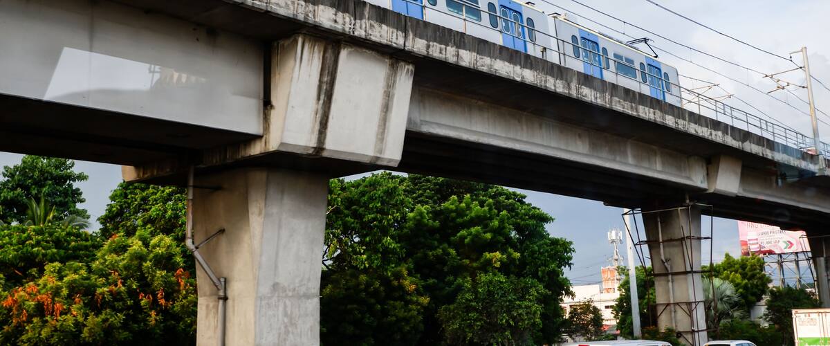 A train on an elevated track with concrete pillars, greenery below, and a billboard in the background.