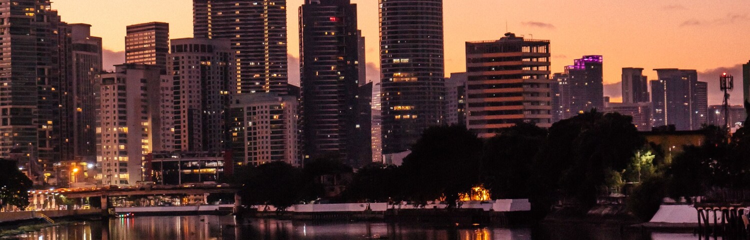 Silhueta urbana ao entardecer, com reflexos em um rio calmo.