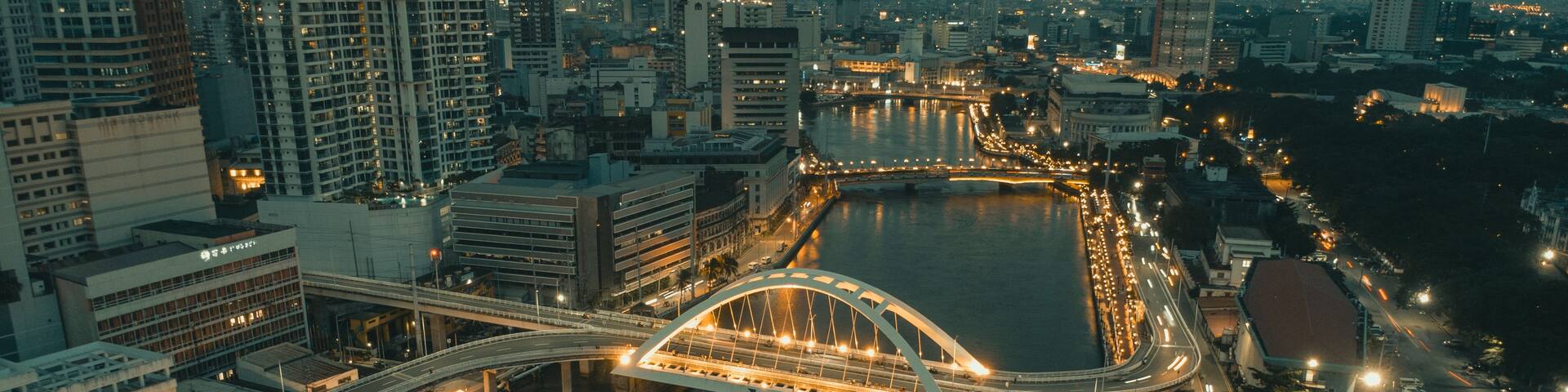 A cityscape at night with a prominent bridge over a river, surrounded by high-rise buildings and illuminated streets.