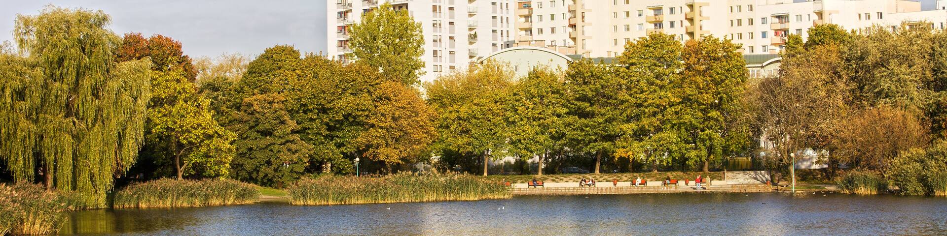 A lake with reflections, surrounded by trees and a row of white apartment buildings.
