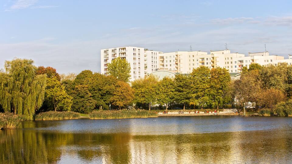 A lake with reflections, surrounded by trees and a row of white apartment buildings.