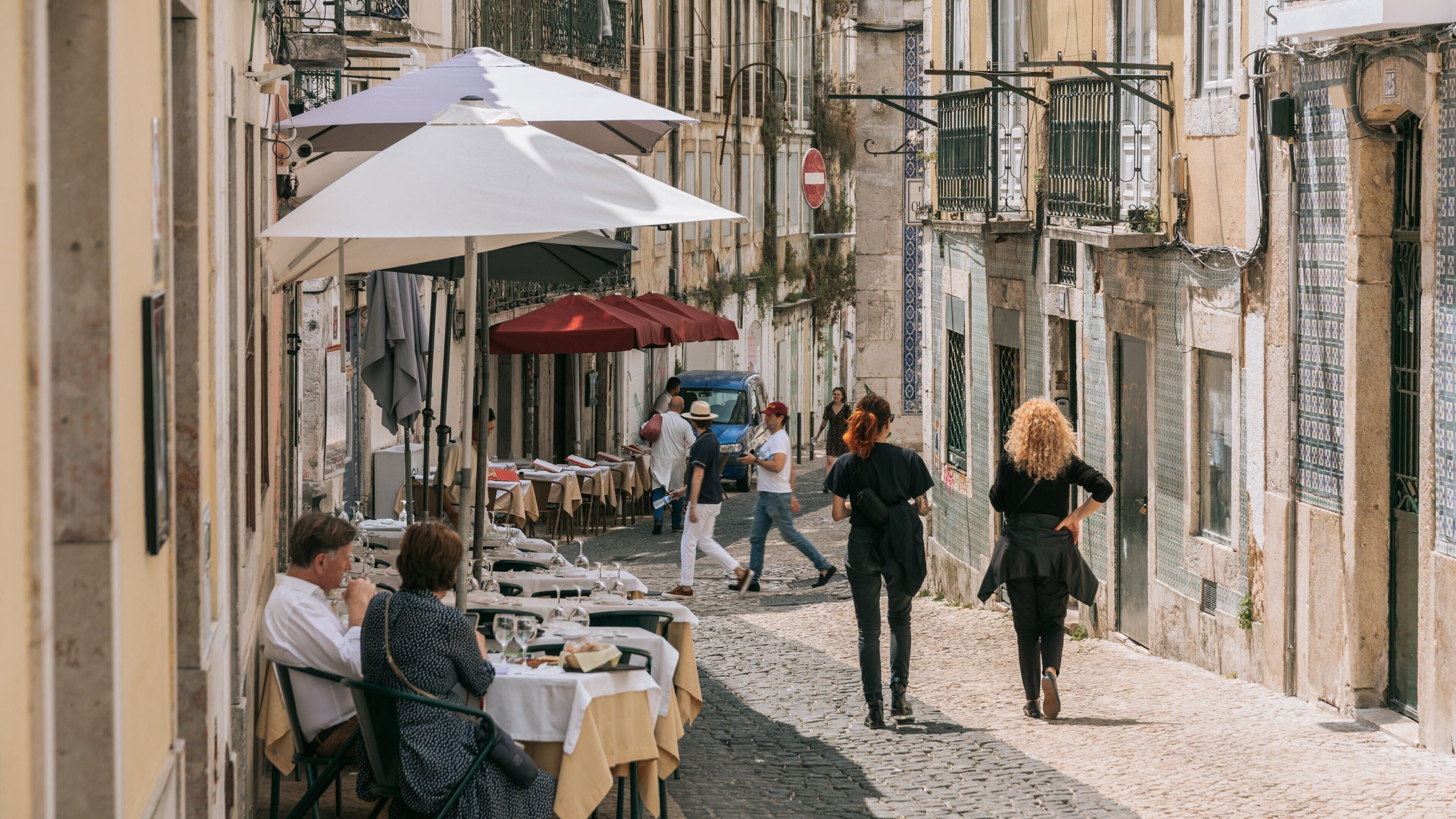 A cobblestone street with outdoor cafes, people walking, and buildings with decorative windows.