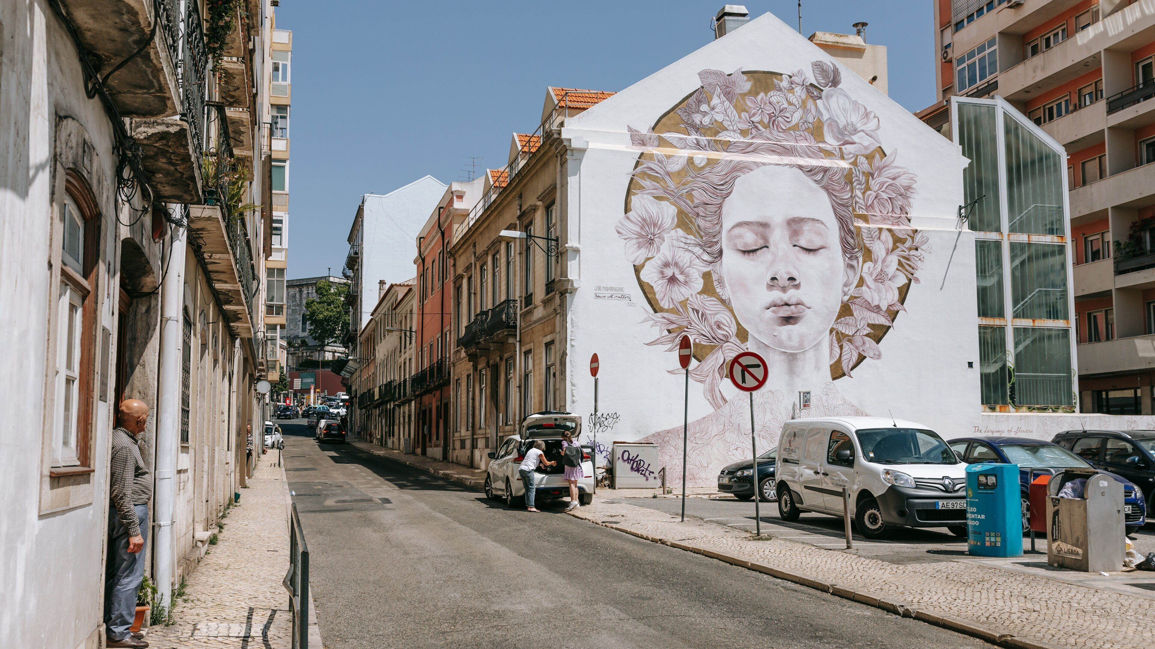 Una calle con un mural grande en un edificio, coches estacionados y una acera adoquinada.