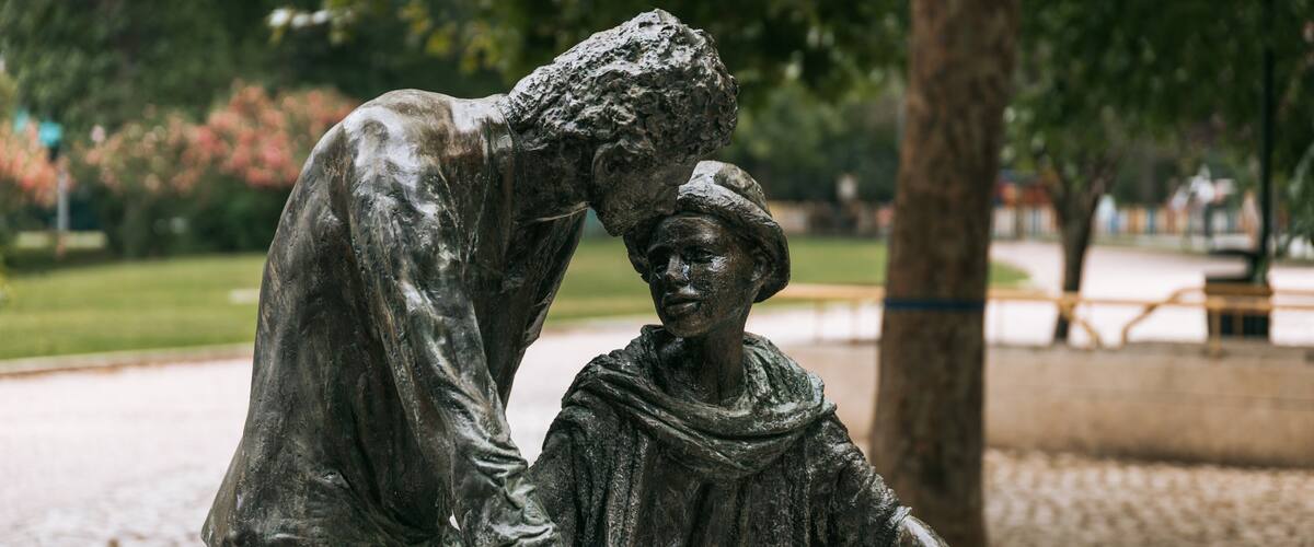 A statue of a person helping another on a bicycle in a park.