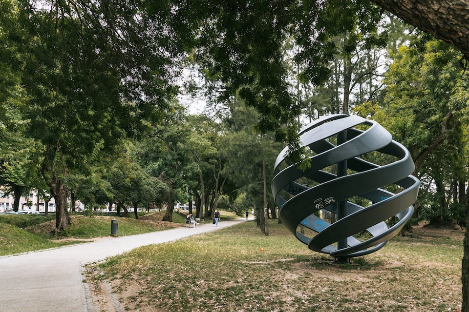 A park with a large metallic sculpture, a paved walkway, and trees.