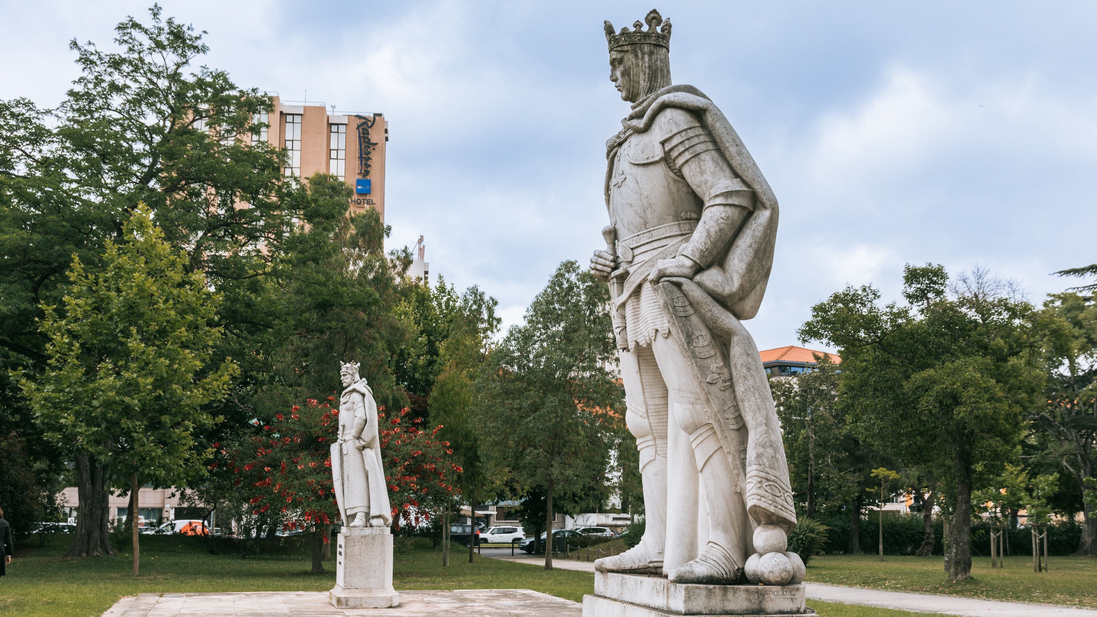 Two statues in a park with trees and a hotel in the background.