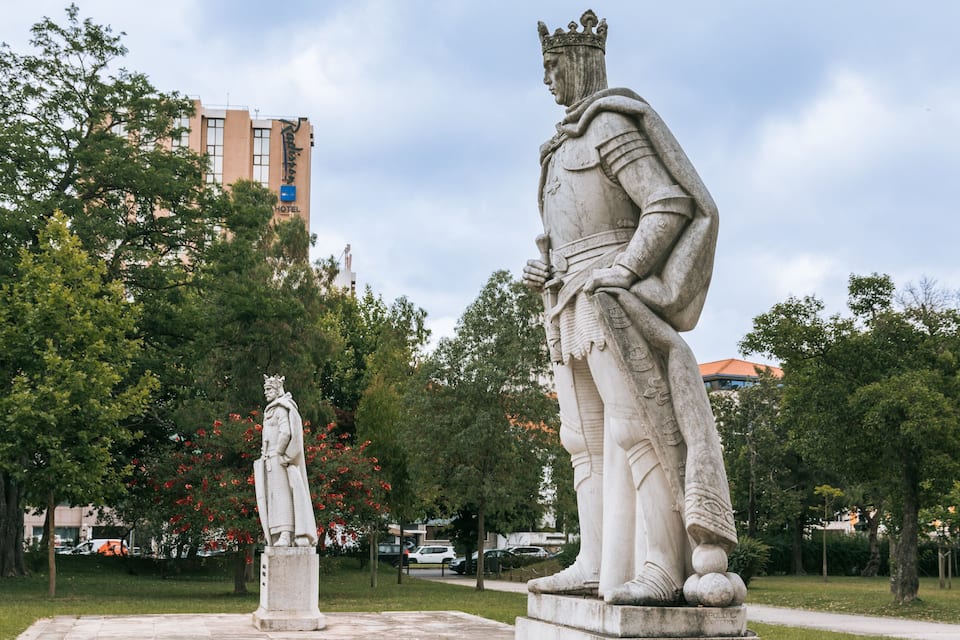 Two statues in a park with trees and a hotel in the background.