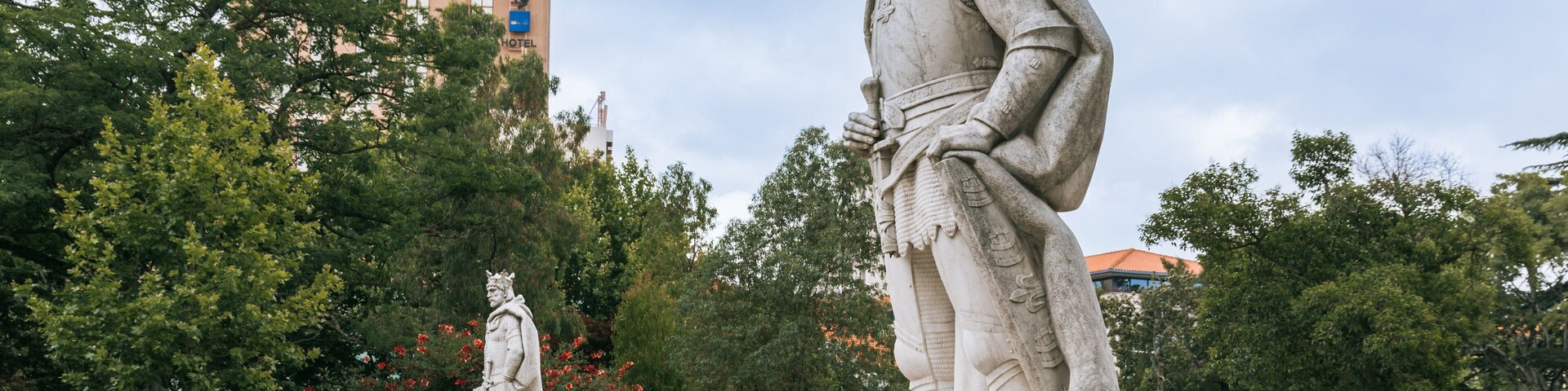 Dos estatuas en un parque con árboles y un hotel al fondo.