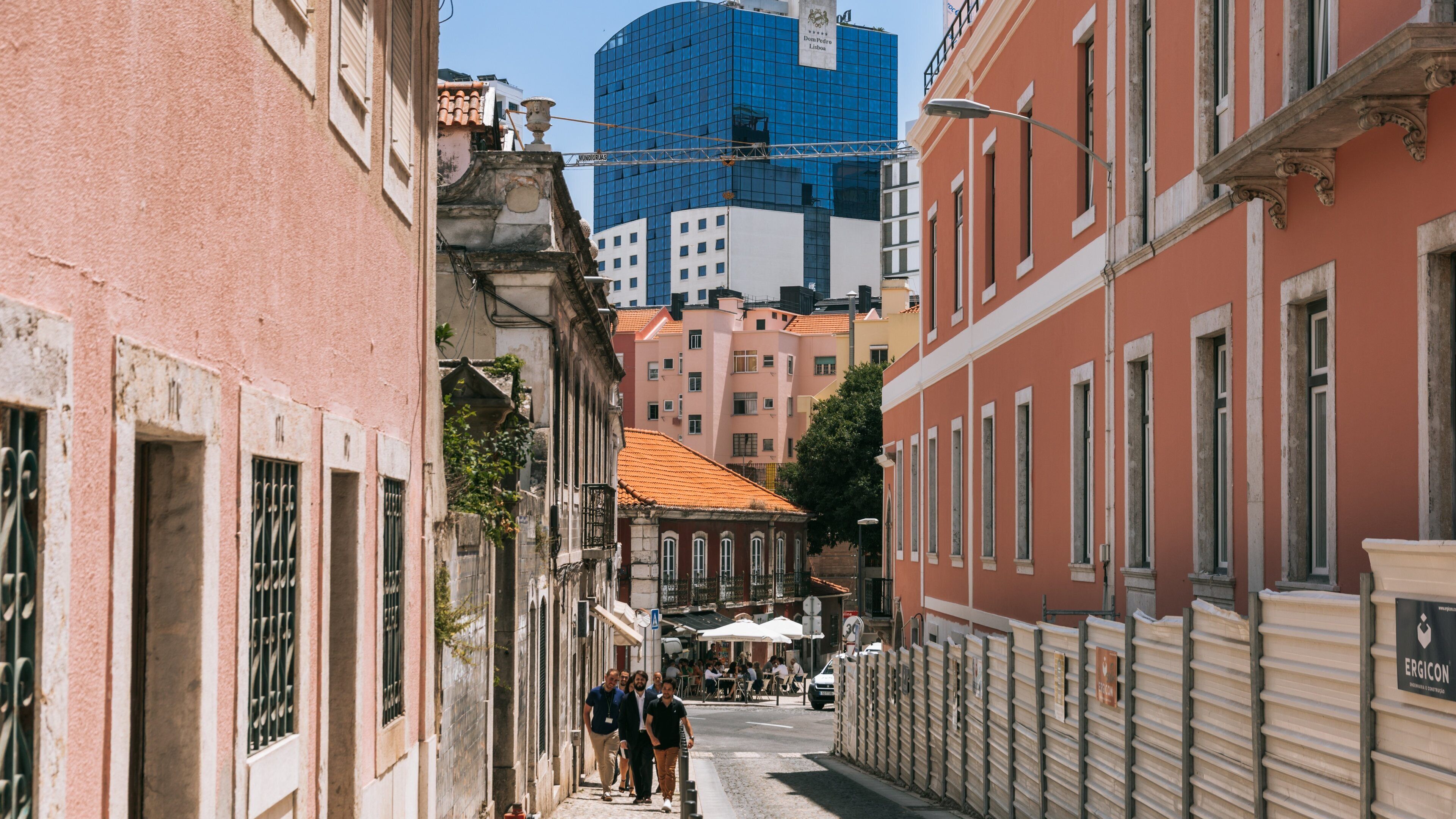 A narrow street lined with historic buildings, a modern blue skyscraper, and outdoor seating at a café.