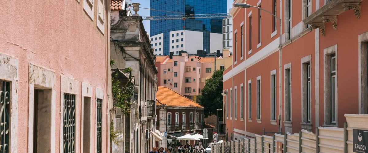 A narrow street lined with historic buildings, a modern blue skyscraper, and outdoor seating at a café.
