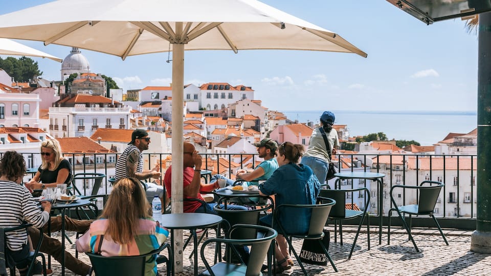 Un groupe de personnes est assis à la terrasse d’un café, profitant d’une vue sur une ville historique et la mer.