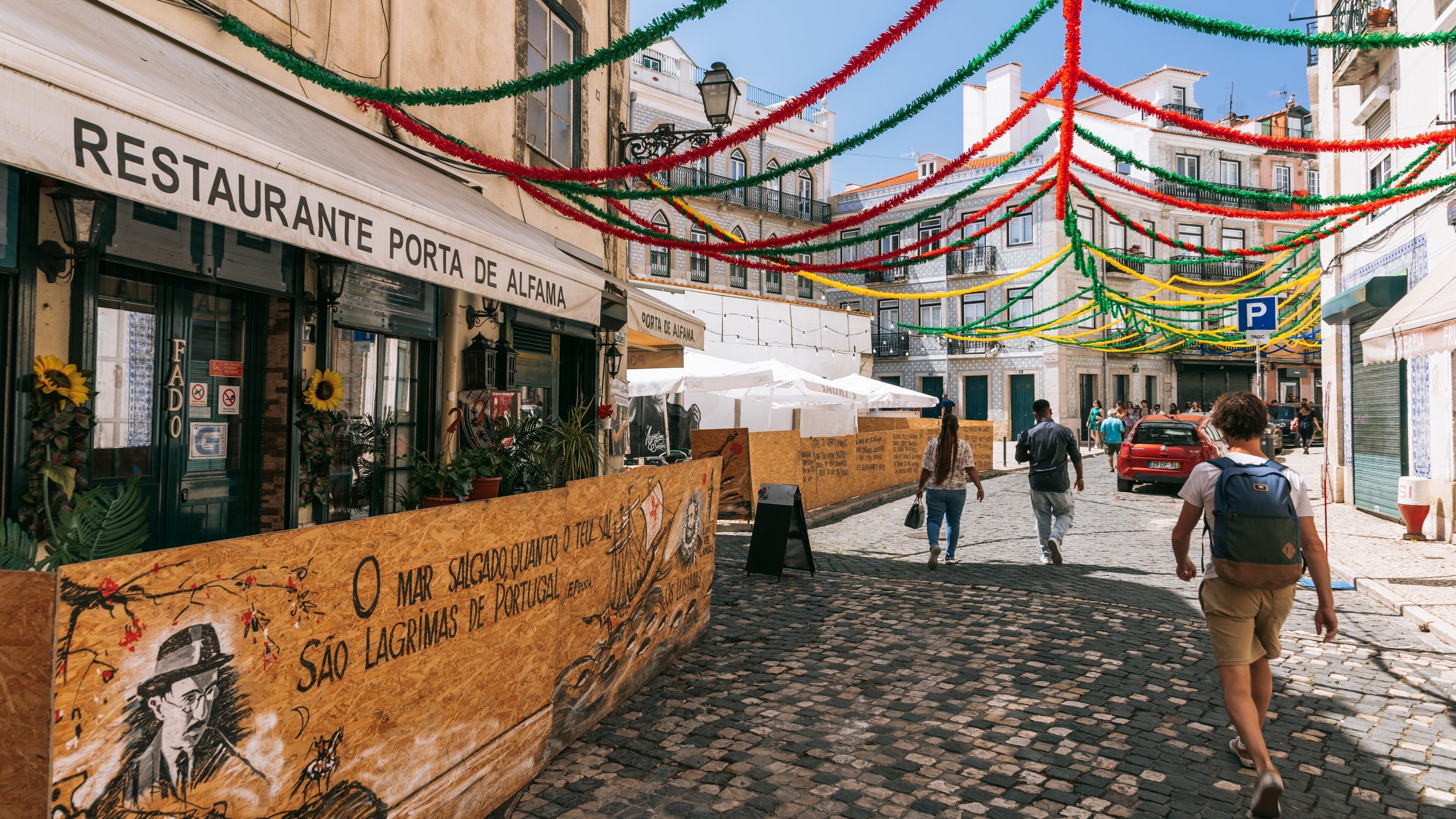 A cobblestone street with a restaurant named 'RESTAURANTE PORTA DE ALEFAMA' and a wooden board with graffiti.