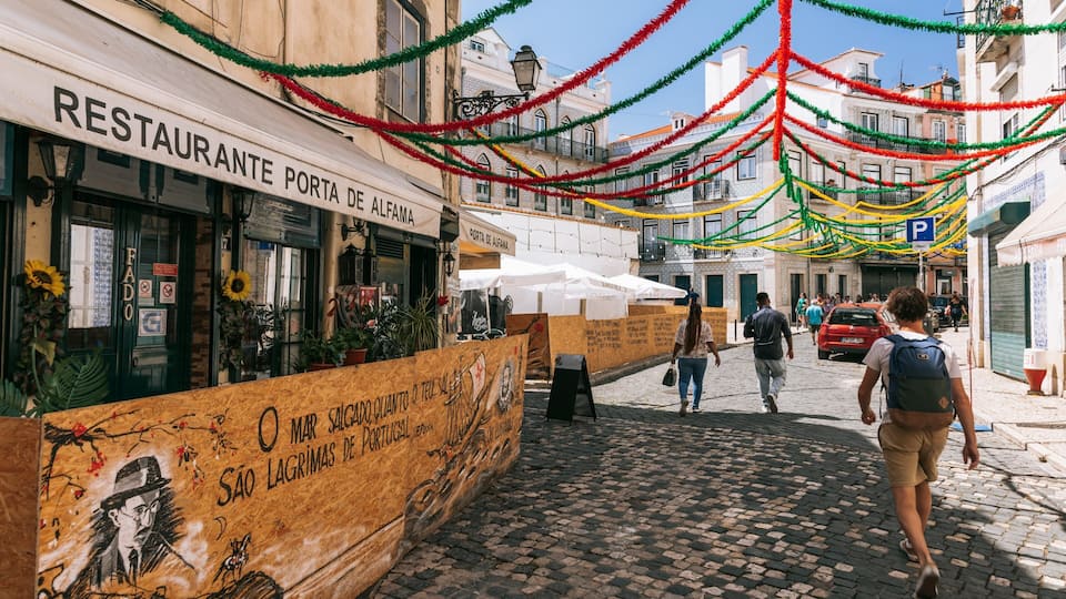 A cobblestone street with a restaurant named 'RESTAURANTE PORTA DE ALEFAMA' and a wooden board with graffiti.
