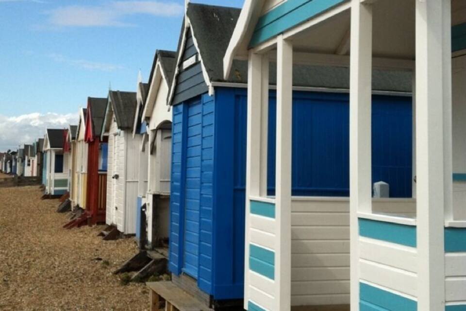 A row of beach huts with blue and white exteriors, numbered sequentially, along a gravel beach.