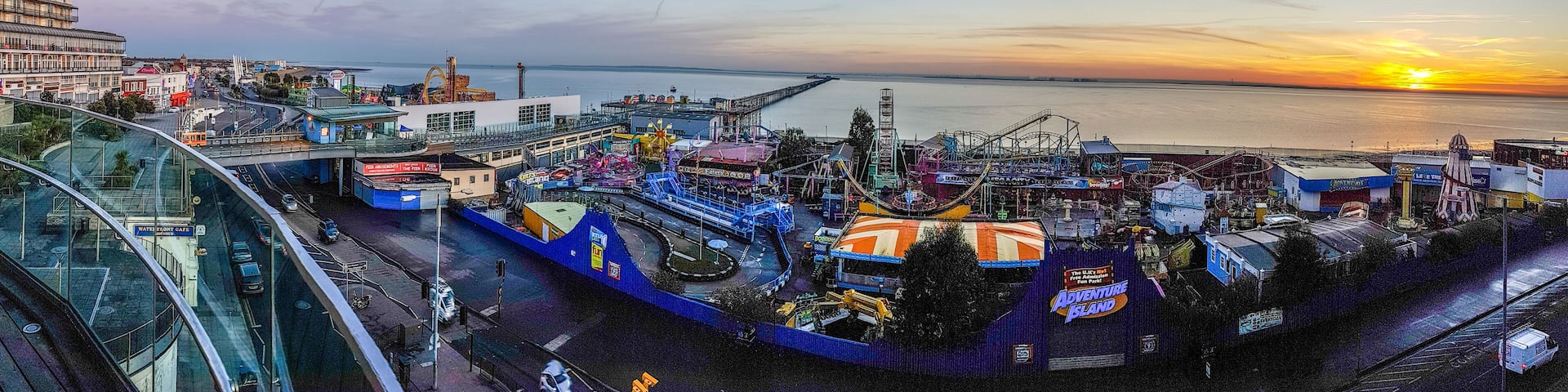 A coastal amusement park at sunset with a roller coaster, Ferris wheel, and a bridge in the background.
