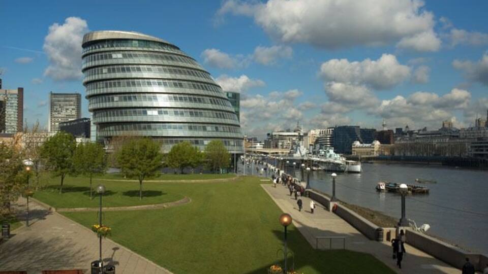 A curved modern building, a river, and a promenade with people walking.