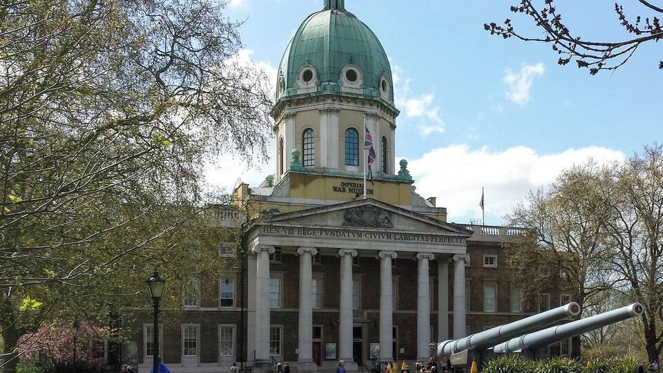 A large building with a green dome and columns, surrounded by trees and a large cannon.