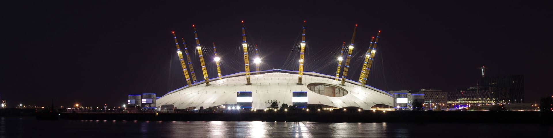 A large, illuminated stadium with a unique, curved roof structure and multiple tall support towers.