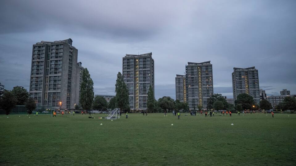 A grassy field with soccer goals and players, surrounded by tall residential buildings.