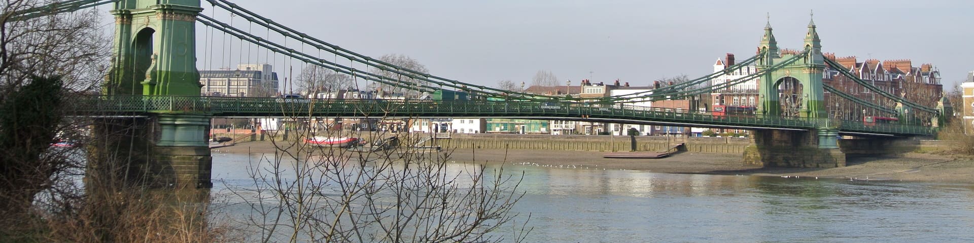 A green suspension bridge over a river with buildings in the background.