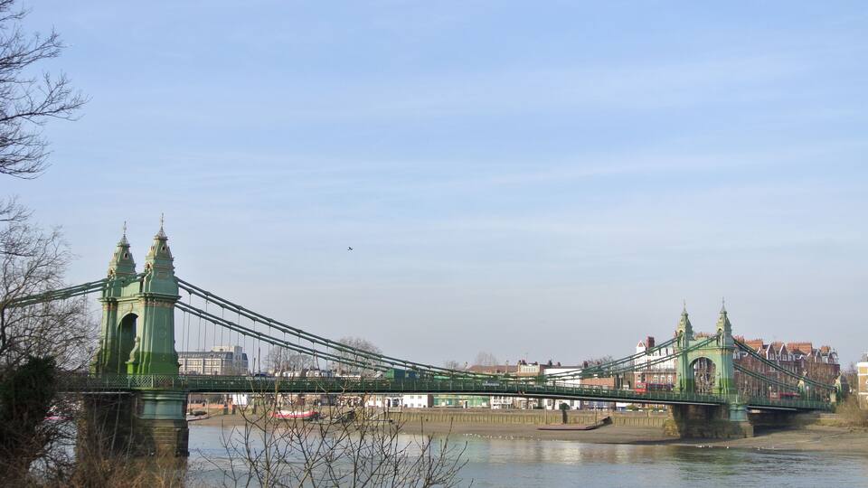 A green suspension bridge over a river with buildings in the background.