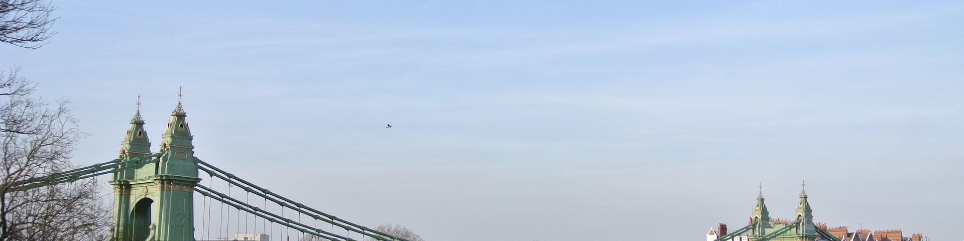 Uma ponte suspensa verde sobre um rio, com edifícios ao fundo.