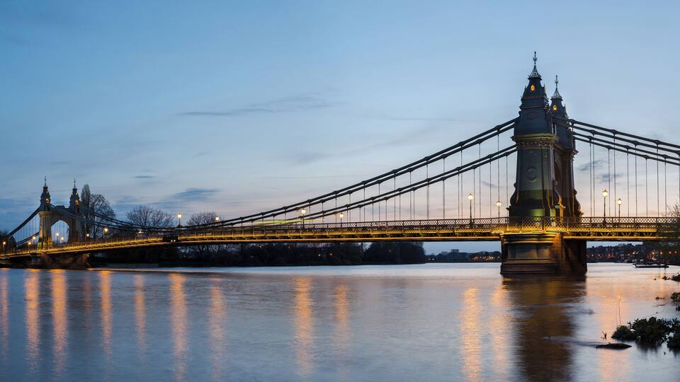 A suspension bridge at dusk with its lights reflecting on the water.